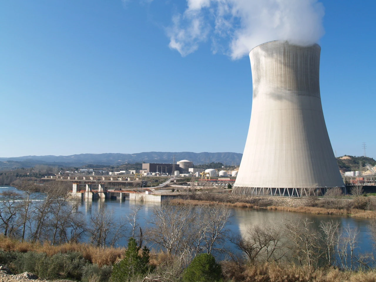 Planta nuclear con torre de refrigeración emitiendo vapor, situada junto a un río y montañas al fondo