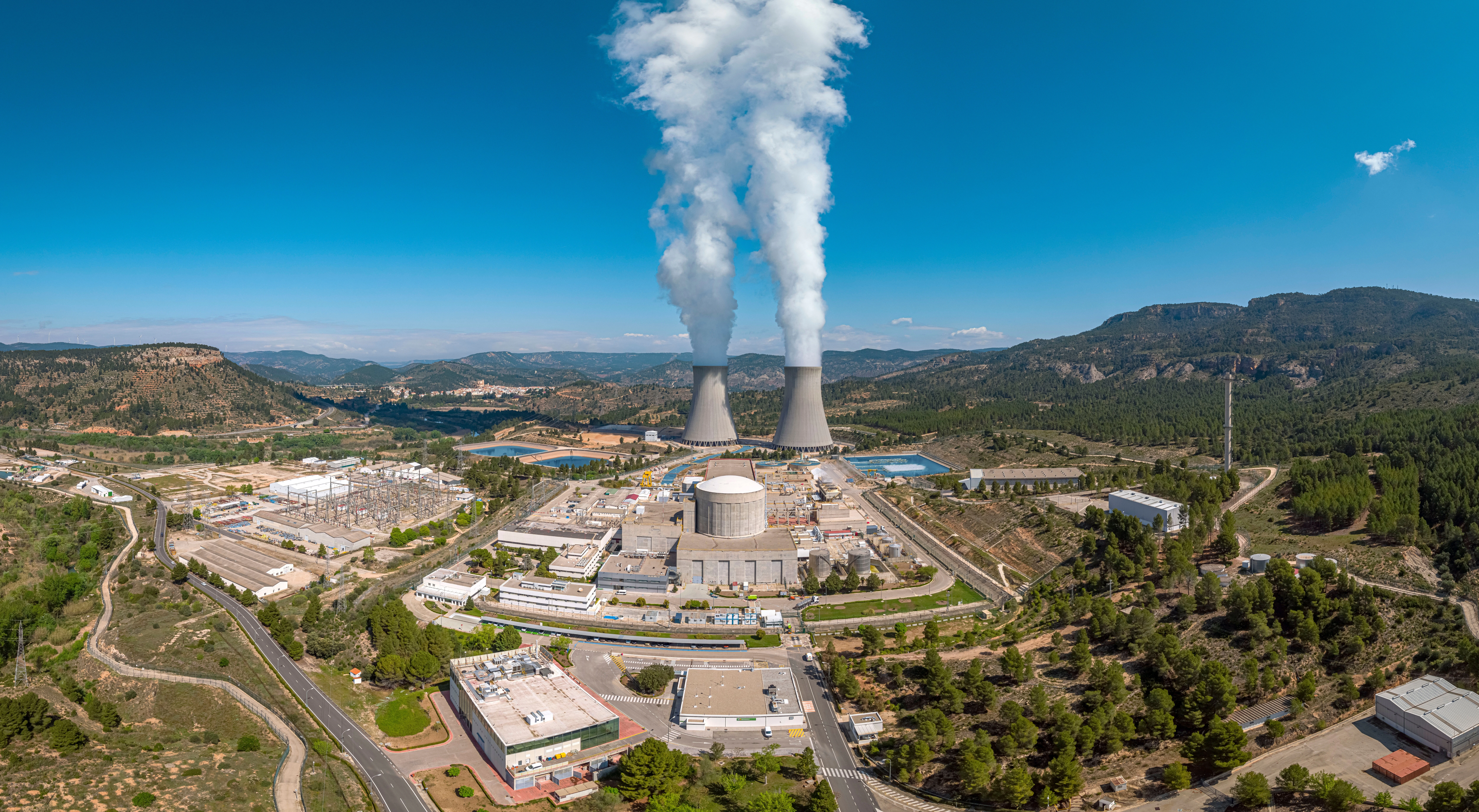 Vista aérea de una planta nuclear con torres de refrigeración emitiendo vapor, rodeada de árboles y montañas