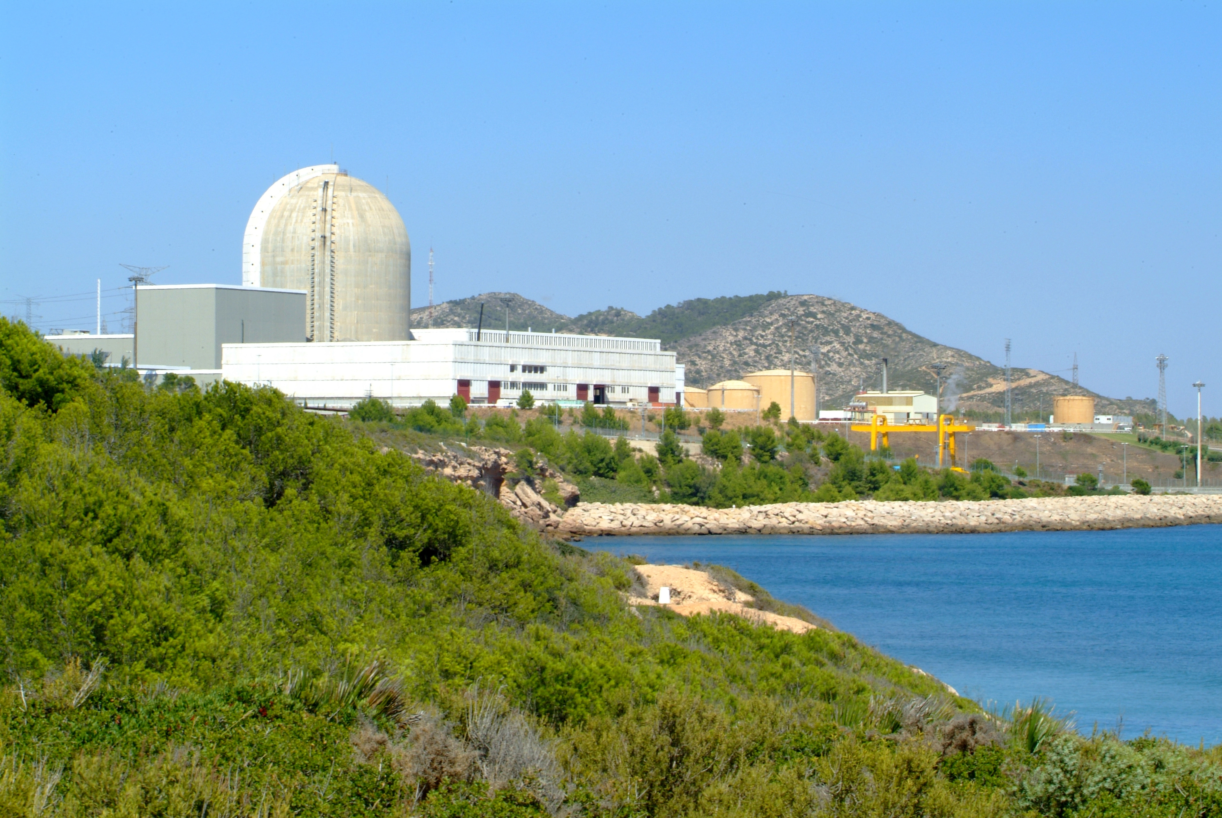 Planta nuclear situada junto al mar, con la torre de refrigeración visible y montañas al fondo