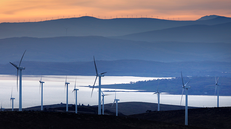 Parque eólico con varios aerogeneradores sobre colinas, cerca de un embalse, y una cadena montañosa al fondo, durante el atardecer.