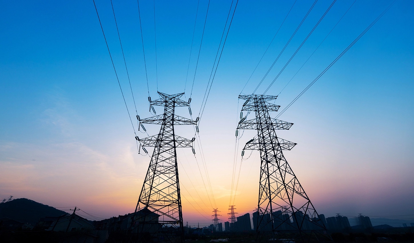 Dos grandes torres de alta tensión en el centro bajo un cielo de atardecer con tonos azules y anaranjados. El sol se pone en el horizonte detrás de las estructuras, y en la base se ven las siluetas de una zona residencial