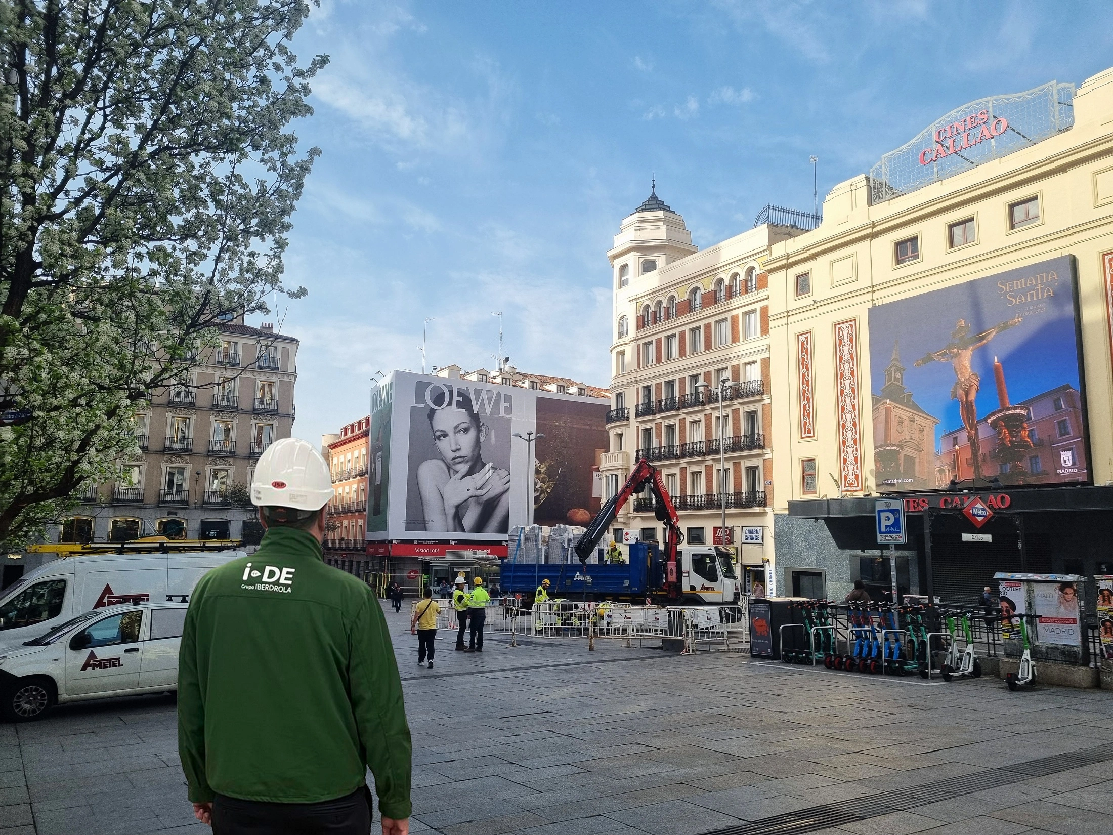 Fotografía de un trabajador de i-DE con casco de seguridad frente a una obra en la Plaza Callao de Madrid, con carteles publicitarios y el edificio de los Cines Callao al fondo.