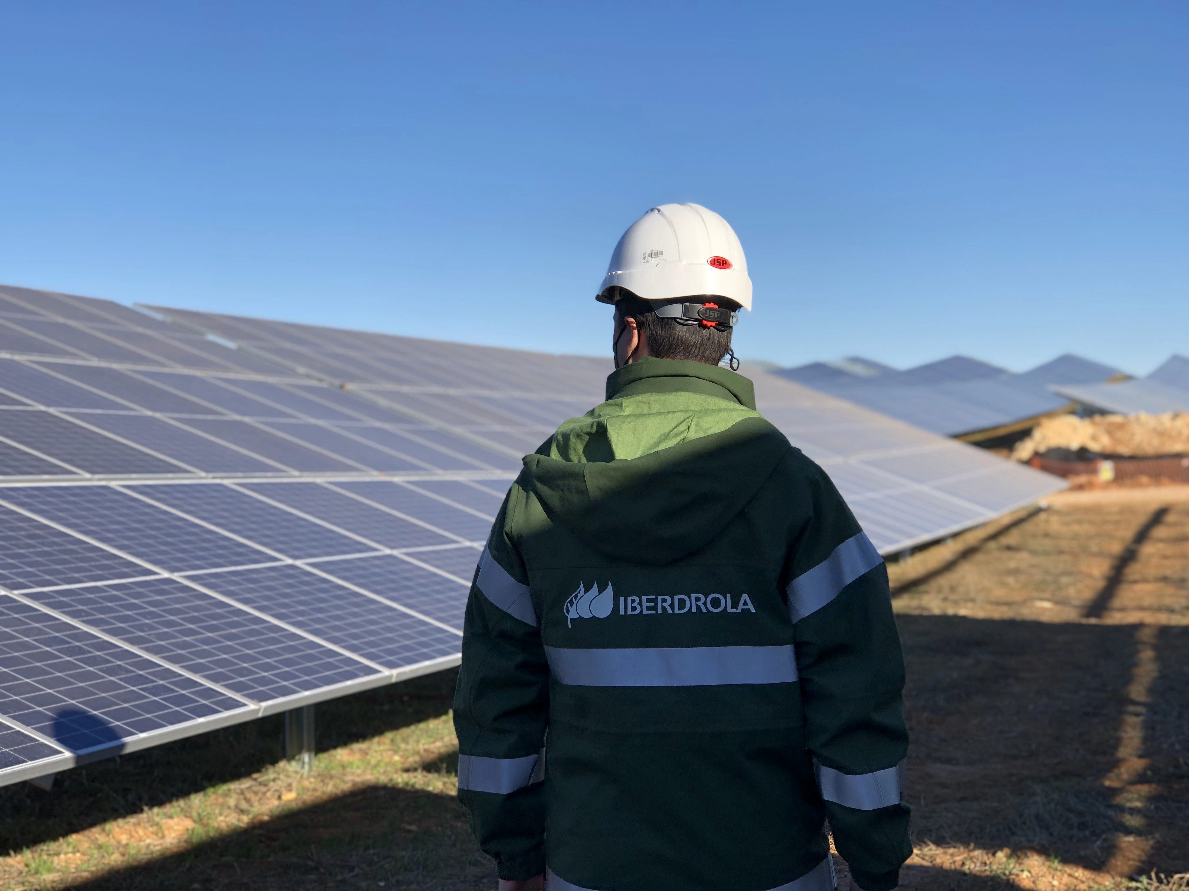 Un trabajador de Iberdrola, con un casco de seguridad y un abrigo verde con franjas reflectantes, está de espaldas frente a un campo de paneles solares. El cielo es claro y se observan montañas a lo lejos. La imagen transmite un enfoque en las energías renovables y el trabajo en una planta fotovoltaica.