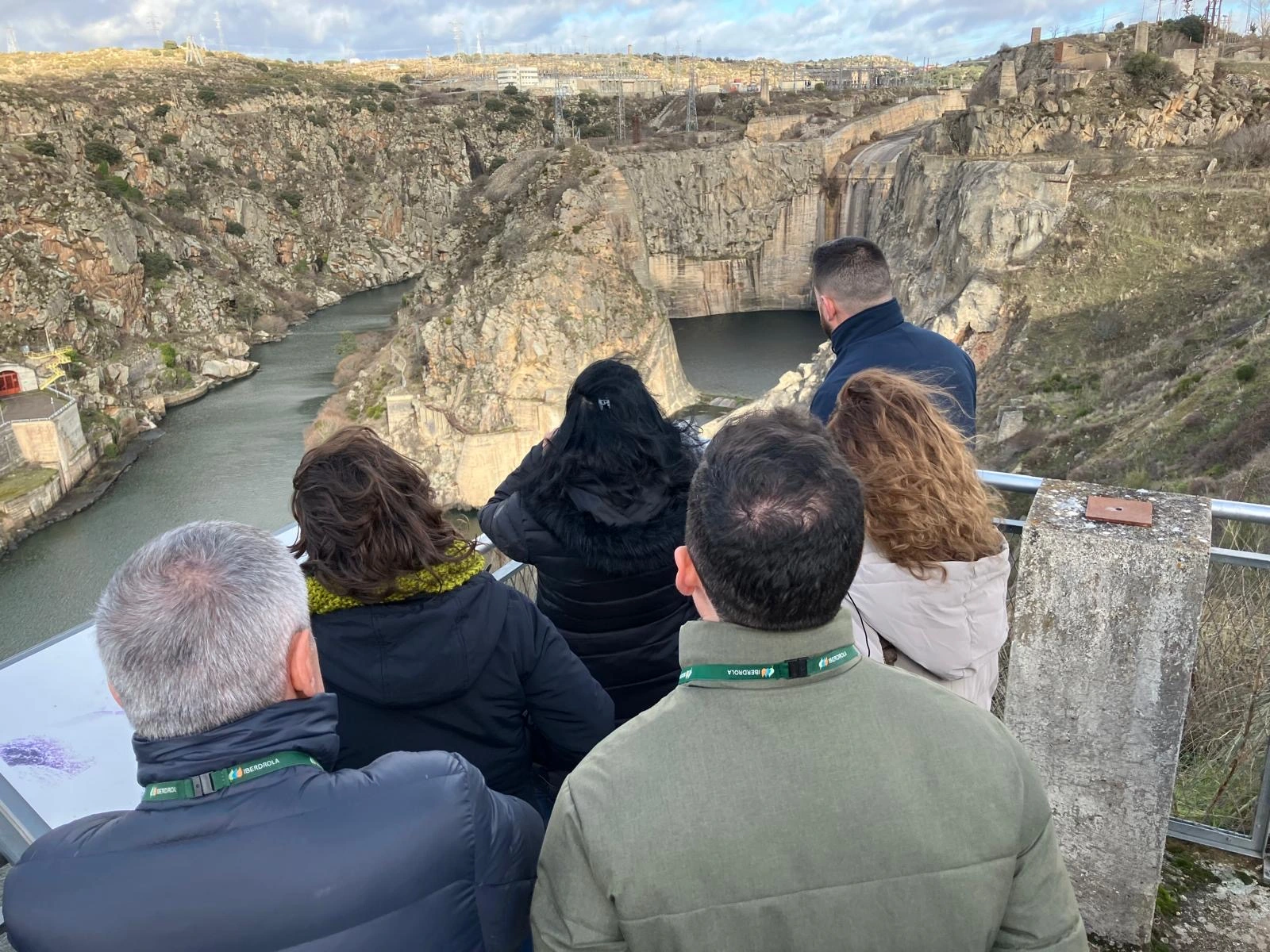 A group of people are observing a rocky landscape and a hydraulic installation from an overlook. They are facing away from the camera, and the scene shows a gorge surrounded by mountains and vegetation. The people are wearing coats, and some have the Iberdrola logo visible on their clothing. In the background, energy structures and power lines are visible.