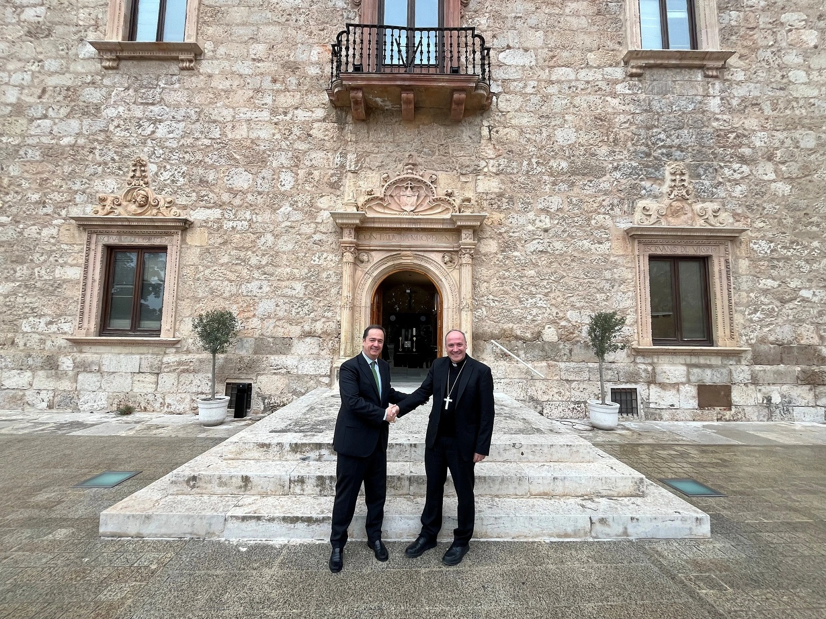 Two people are shaking hands in front of an entrance to an old building. The man on the left is wearing a dark suit with a green tie, while the man on the right is wearing a black cassock with a visible crucifix. Both are standing on the stone staircase leading to the main door of the building, which has a stone façade with architectural details