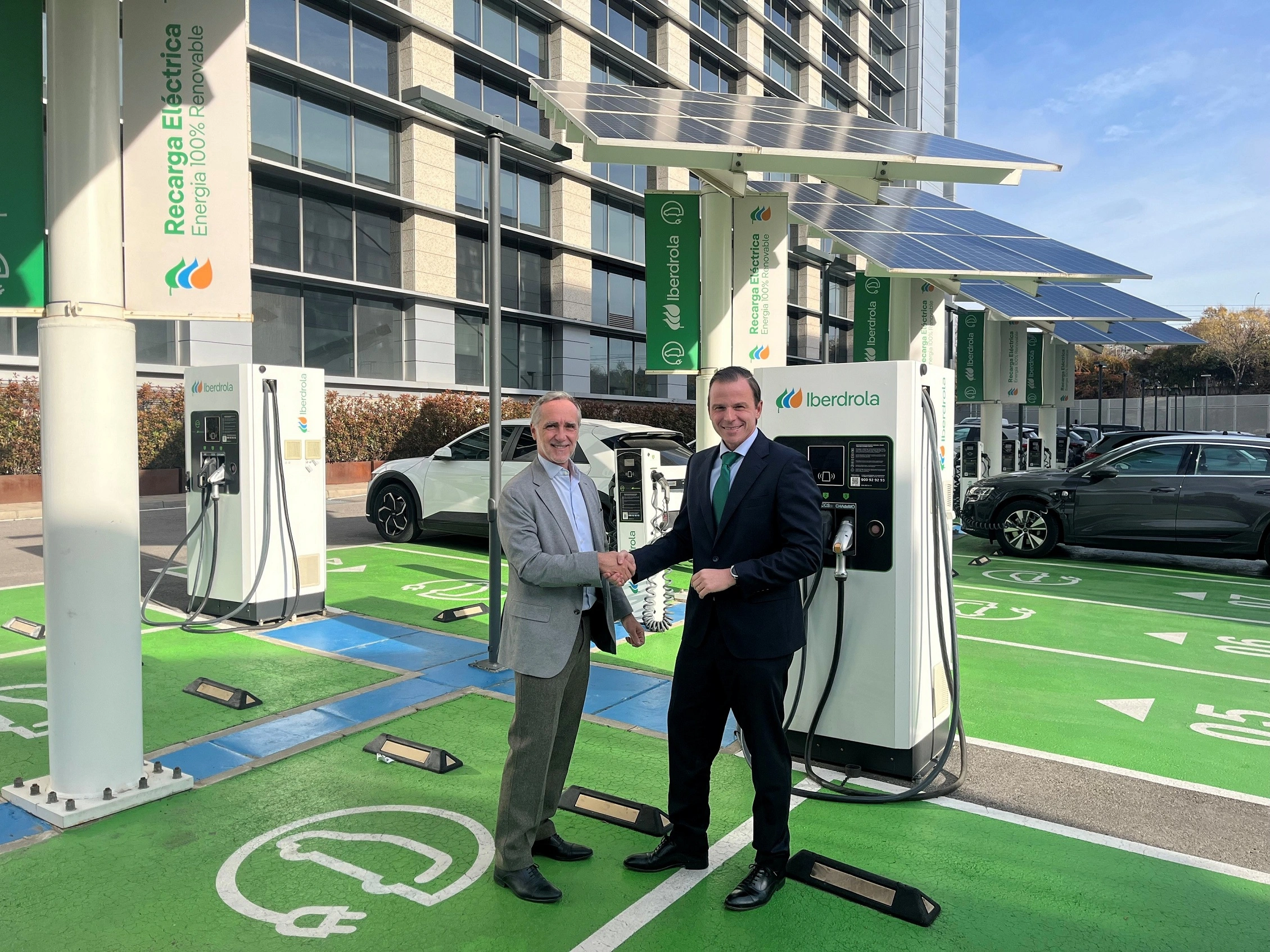 Two men are shaking hands in front of an electric vehicle charging station, which is equipped with solar panels. Both are standing next to one of the chargers, with electric cars parked in the designated spaces. The chargers have the Iberdrola logo, and in the background, a modern building with large windows can be seen. The sign reads "Electric Charging, 100% Renewable Energy.