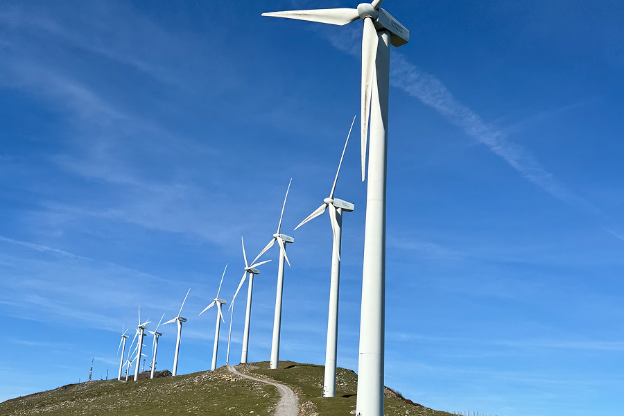 Vista de un parque eólico con varias turbinas de viento alineadas en una colina. El cielo es claro y azul, con algunas nubes dispersas. Un camino serpenteante en primer plano conduce hacia las turbinas.