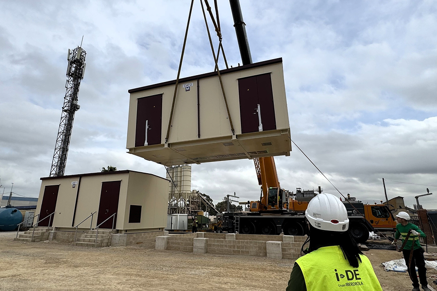 En la imagen, un contenedor de gran tamaño está siendo levantado por una grúa en una obra de construcción. El contenedor, de color beige con puertas rojas, es transportado sobre un terreno con estructura de cemento. En primer plano, se puede ver a un trabajador con chaleco y casco de seguridad de Iberdrola, observando el proceso.