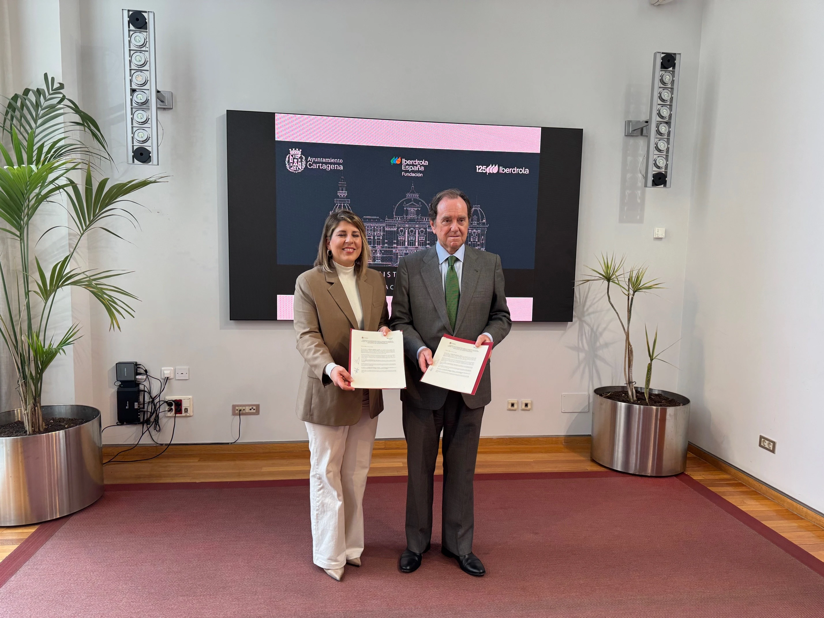 Una mujer y un hombre posan juntos mientras sostienen documentos firmados en una ceremonia, con el fondo de una pantalla que muestra el logo de Iberdrola y la imagen de un edificio. La mujer viste una chaqueta beige y pantalones claros, y el hombre lleva un traje gris con una corbata verde. En el fondo, se observan plantas decorativas y una pared moderna de oficina.