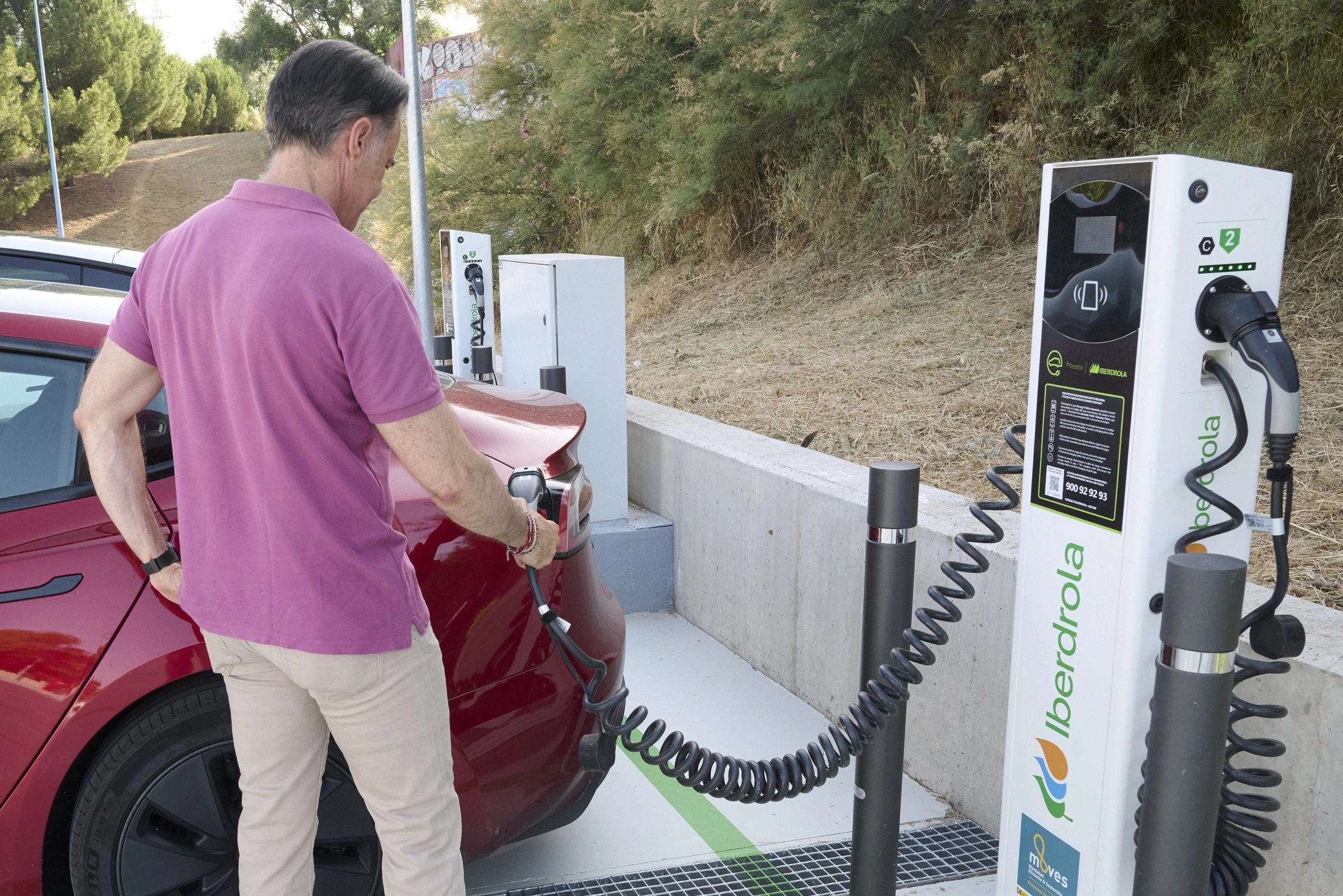 Un conductor está cargando su coche eléctrico en un punto de recarga de Iberdrola. La estación de carga está ubicada al aire libre, y se puede ver claramente el logo de Iberdrola en la estación de carga. El conductor, de espaldas, sostiene el cable de carga y lo conecta al coche rojo. El fondo muestra vegetación y una estructura simple de cemento.