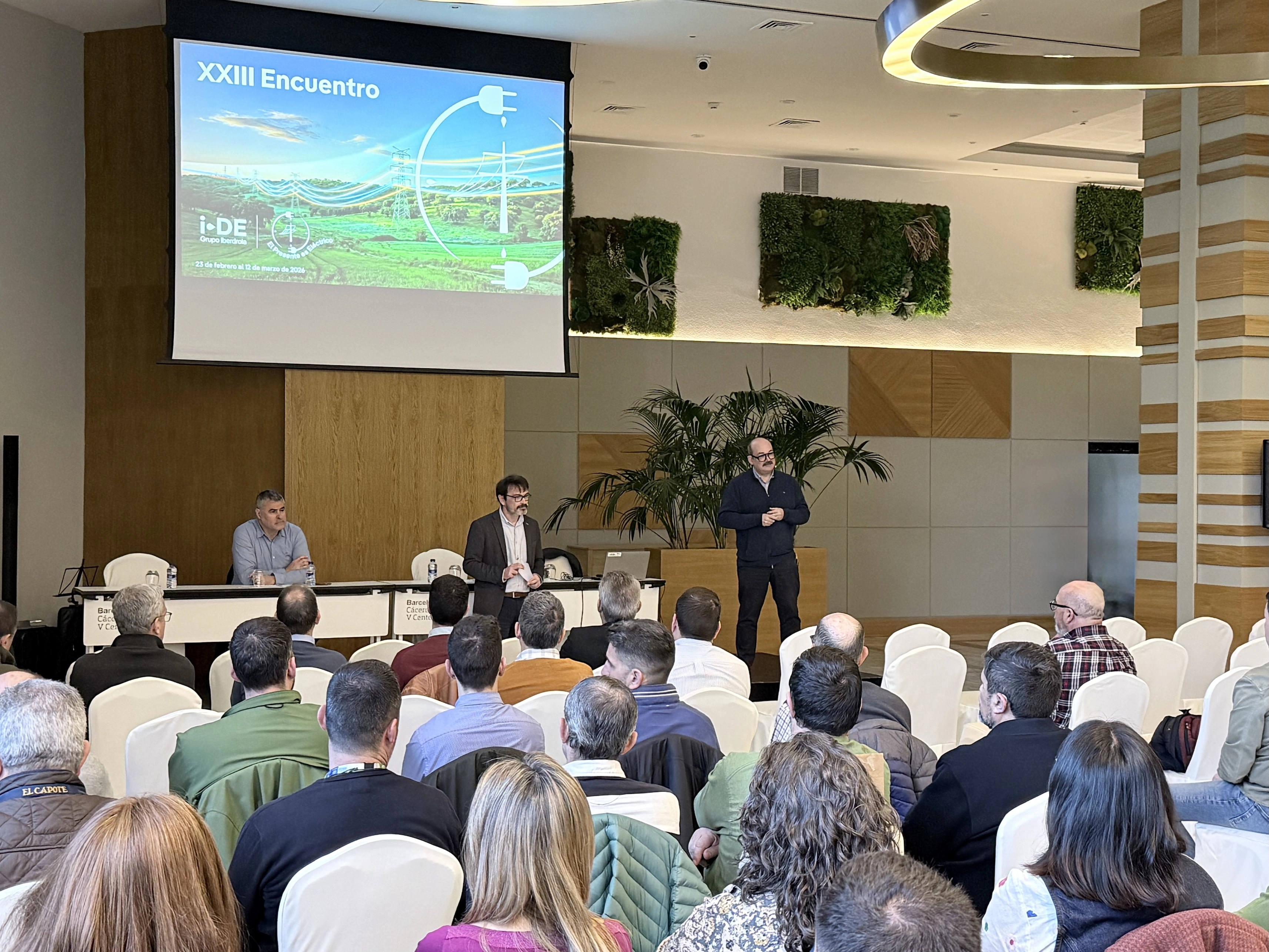 Sala de conferencias con público asistiendo a una presentación. En el escenario, tres hombres participan en una mesa redonda: dos sentados tras una mesa y uno de pie hablando al público. En la pantalla proyectada se lee “XXIII Encuentro” junto al logotipo de i-DE Grupo Iberdrola y una imagen relacionada con redes eléctricas. La sala cuenta con decoración moderna y paneles vegetales en la pared.