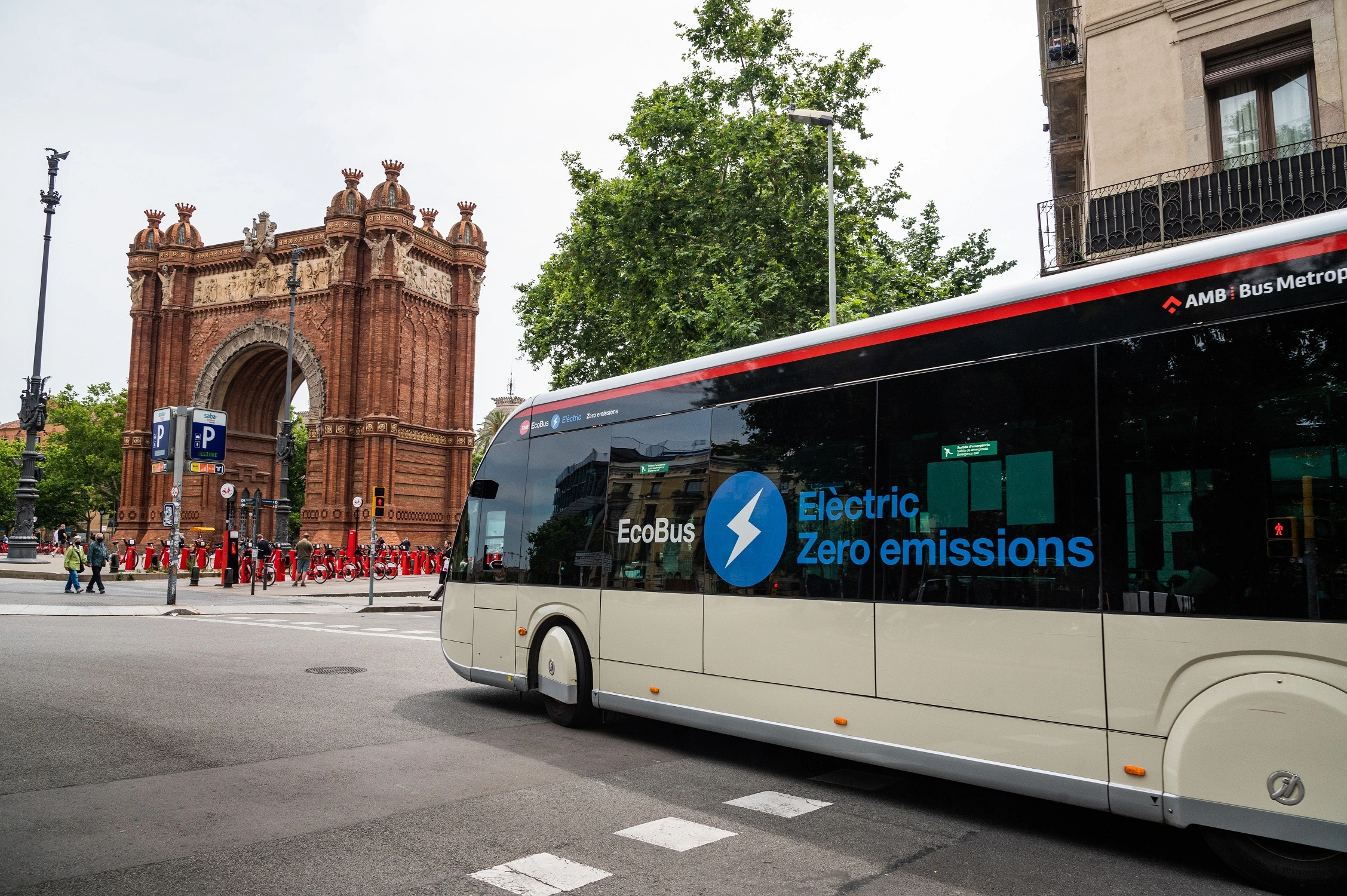 Un autobús eléctrico de TMB (Transports Metropolitans de Barcelona) circula por una calle de la ciudad, pasando frente al Arco del Triunfo. El autobús lleva la marca EcoBus en su lateral, con un logotipo que resalta las palabras "Electric" y "Zero emissions", destacando su naturaleza ecológica. El entorno muestra edificios y árboles, y hay una fila de personas en la acera.