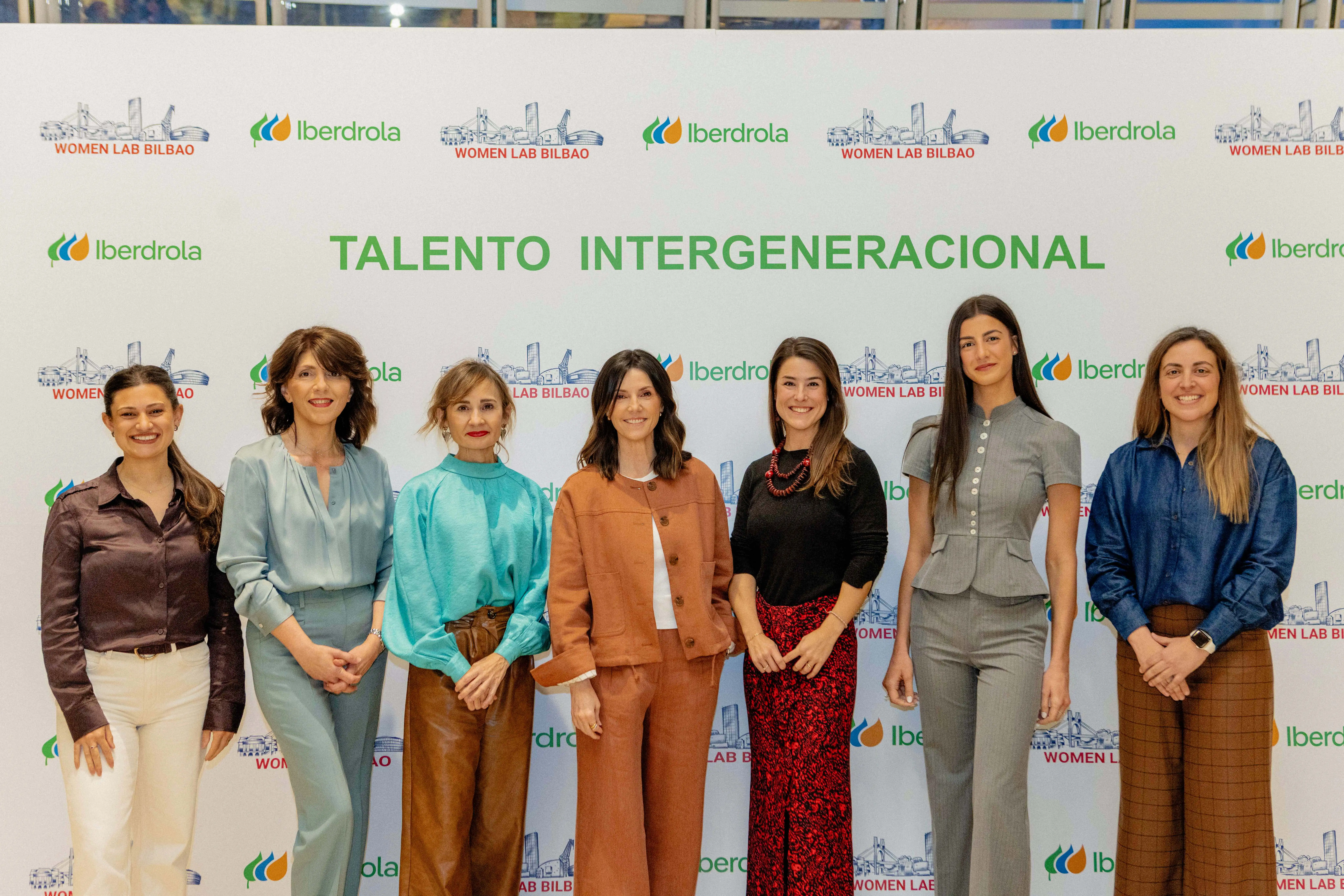 Un grupo de mujeres se encuentra posando frente a un cartel con el logo de Iberdrola y el eslogan "Talento Intergeneracional" en el evento Women Lab Bilbao. Cada mujer lleva una vestimenta moderna y colorida, y todas lucen sonrientes.
