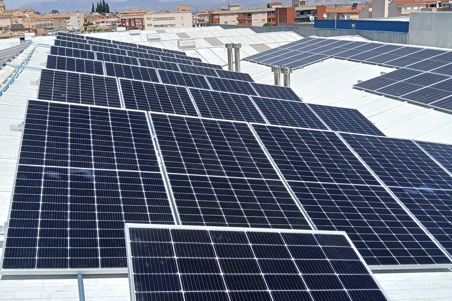Vista aérea de un techo con paneles solares instalados en una comunidad de Granada. Los paneles ocupan una gran parte de la superficie del techo, captando la luz del sol para generar energía renovable. Al fondo, se pueden ver los edificios de la ciudad con una línea de cielo despejado.