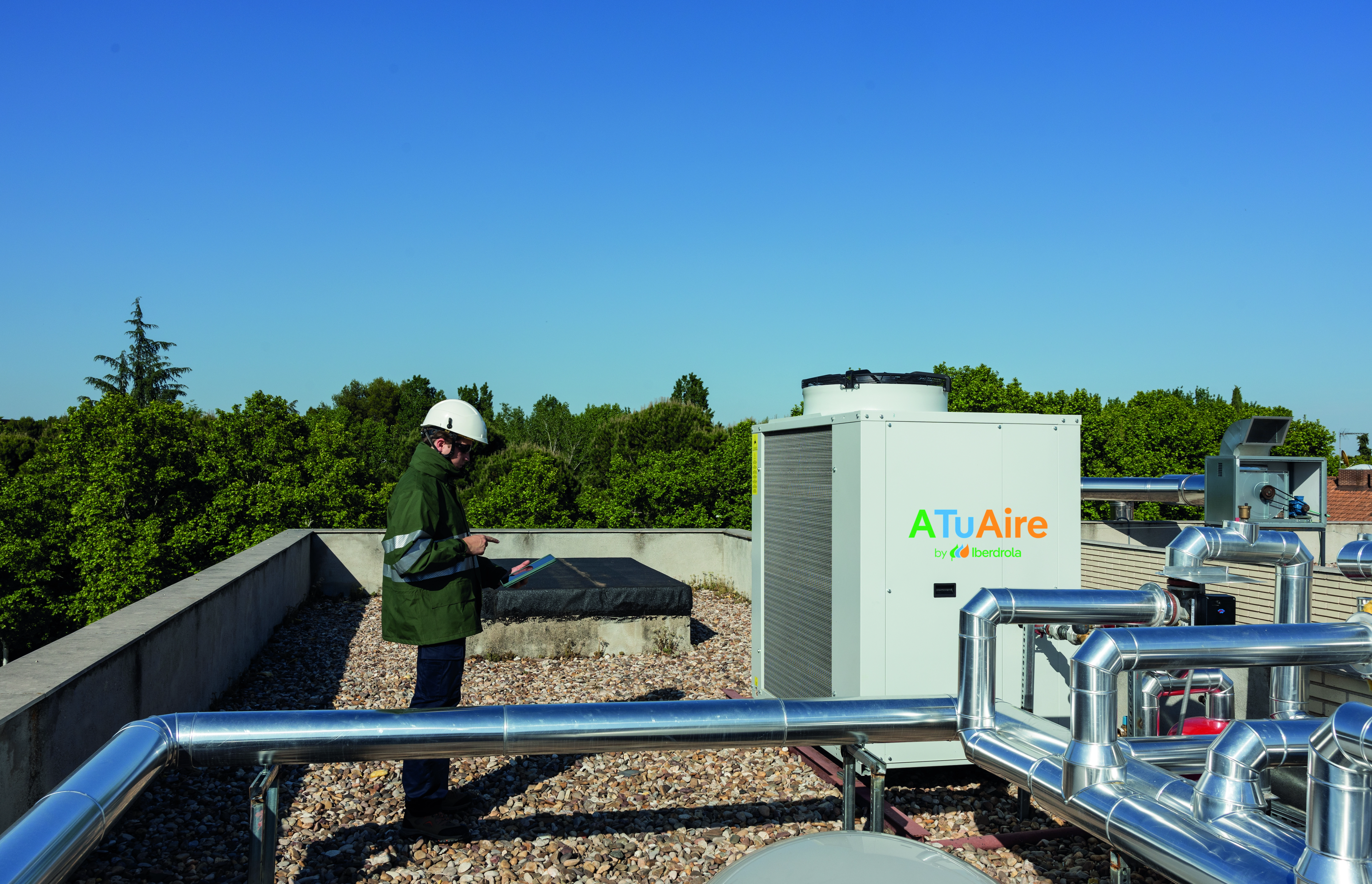 Un trabajador de Iberdrola España con casco de seguridad y abrigo verde, observa una unidad de aerotermia ATuAire en la azotea de un edificio. Al fondo, se aprecian tuberías metálicas y un cielo despejado con árboles.