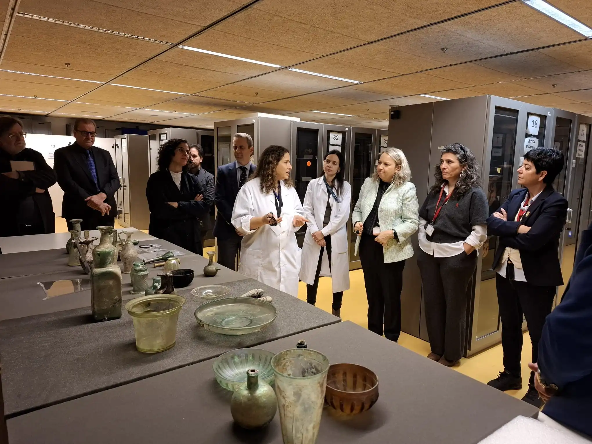 Un grupo de personas en una sala de almacenamiento de un museo, donde se observan objetos de vidrio sobre una mesa. Una mujer, vestida con bata blanca, está explicando el proceso de restauración a los demás, quienes parecen ser colegas o visitantes. La escena muestra varias vitrinas y estanterías con más objetos en el fondo.