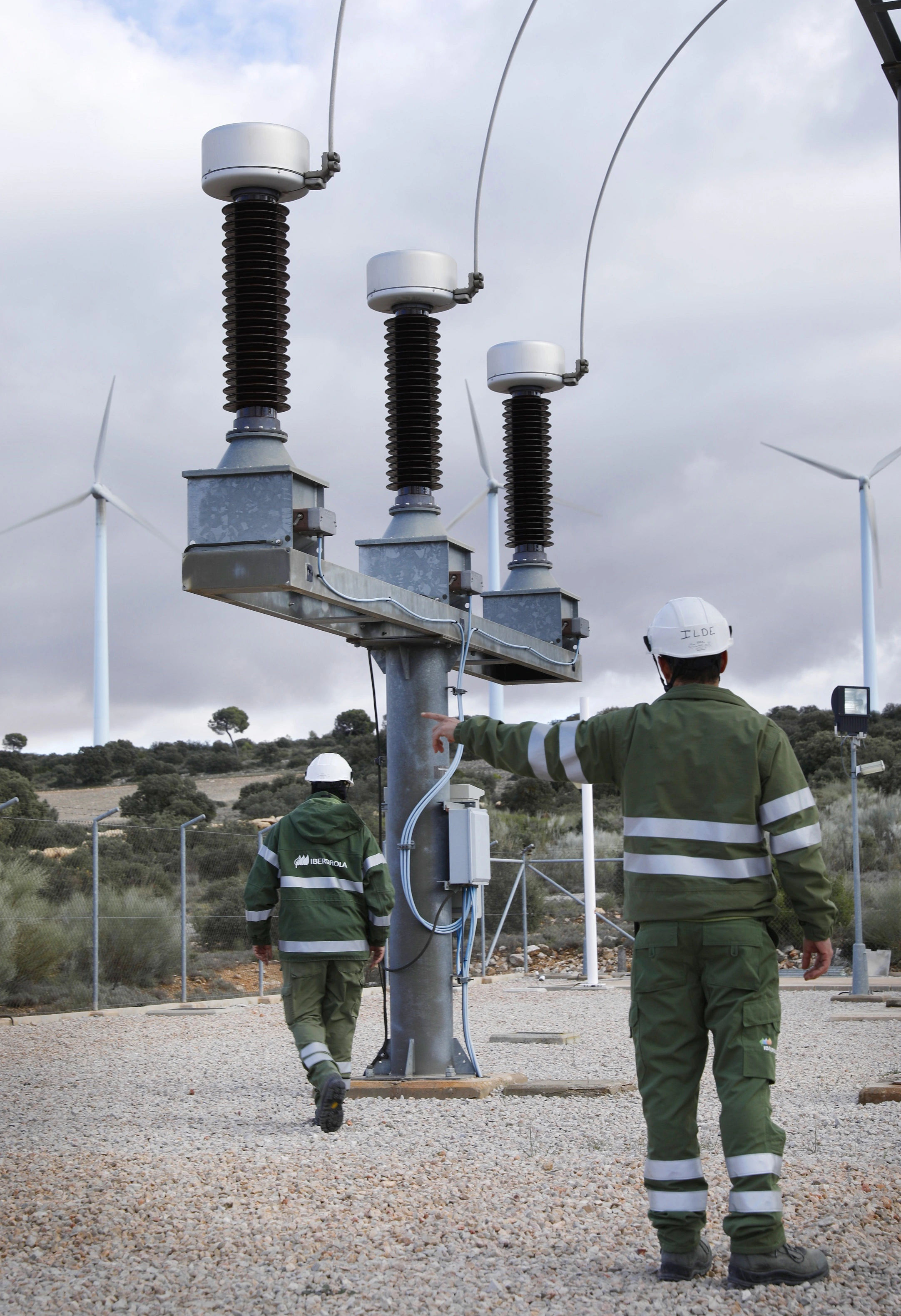 Trabajadores de i-DE en una subestación eléctrica, supervisando equipos y observando aerogeneradores al fondo.