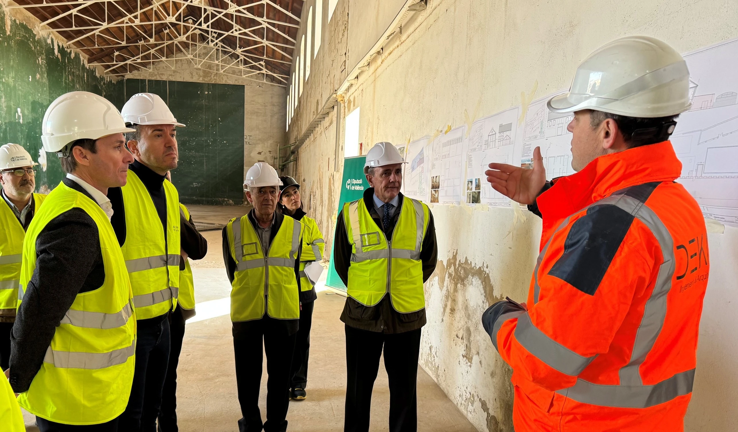 Un grupo de personas, todas con cascos de seguridad y chalecos reflectantes amarillos, escucha atentamente a un hombre con chaqueta naranja reflectante que está explicando algo frente a los planos colgados en la pared. El hombre de pie, con chaleco naranja, parece ser el responsable de la obra. En el fondo, se ve un espacio amplio y sin terminar, con paredes que muestran signos de desgaste. La escena tiene lugar en lo que parece ser una visita o presentación en un lugar de rehabilitación de un edificio.