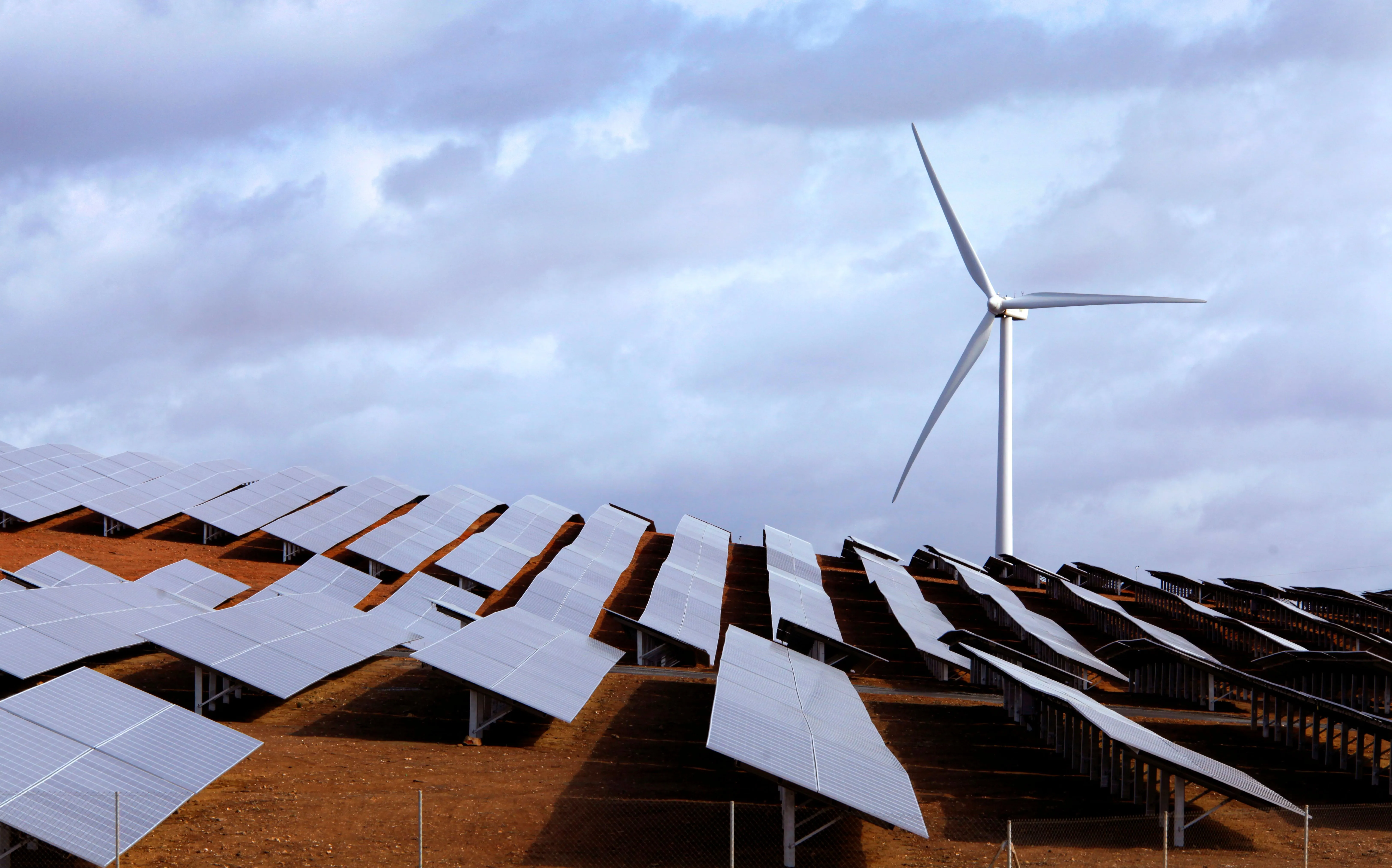 Paneles solares alineados en un terreno rojizo con un aerogenerador al fondo bajo cielo parcialmente nublado en el Parque Andévalo de Iberdrola España.