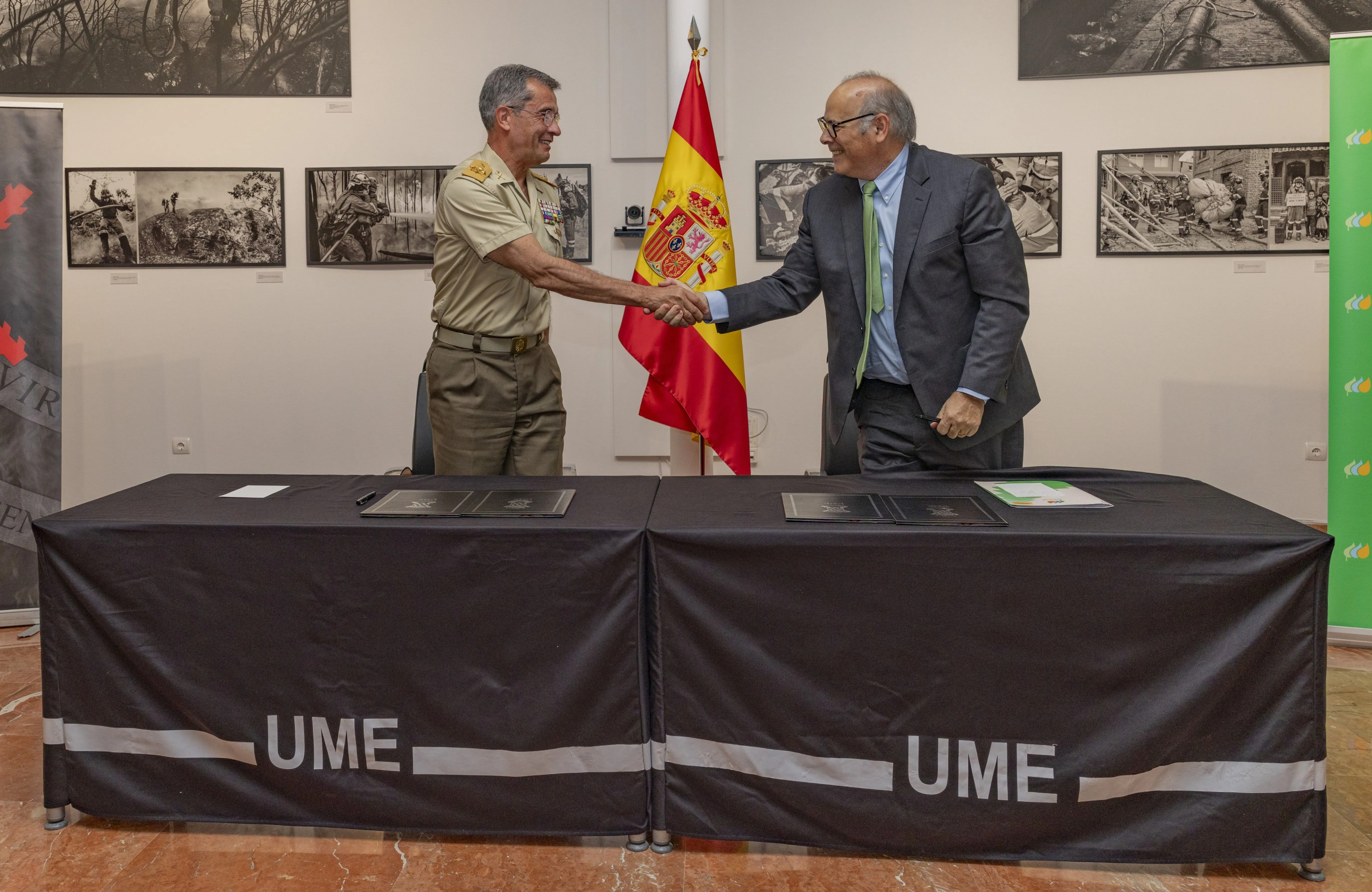 Teniente general Francisco Javier Marcos Izquierdo, Jefe de la UME, y Mario Ruiz-Tagle, CEO de Iberdrola España, se dan la mano frente a una mesa cubierta con manteles negros con el logo de la UME. Detrás se ve la bandera de España, fotografías en blanco y negro en la pared y un roll-up verde con el logo de Iberdrola. Ambos sonríen, vestidos formalmente; Marcos Izquierdo con uniforme militar y Ruiz-Tagle con traje y corbata. En la mesa hay carpetas y bolígrafos, indicando la firma de un convenio.