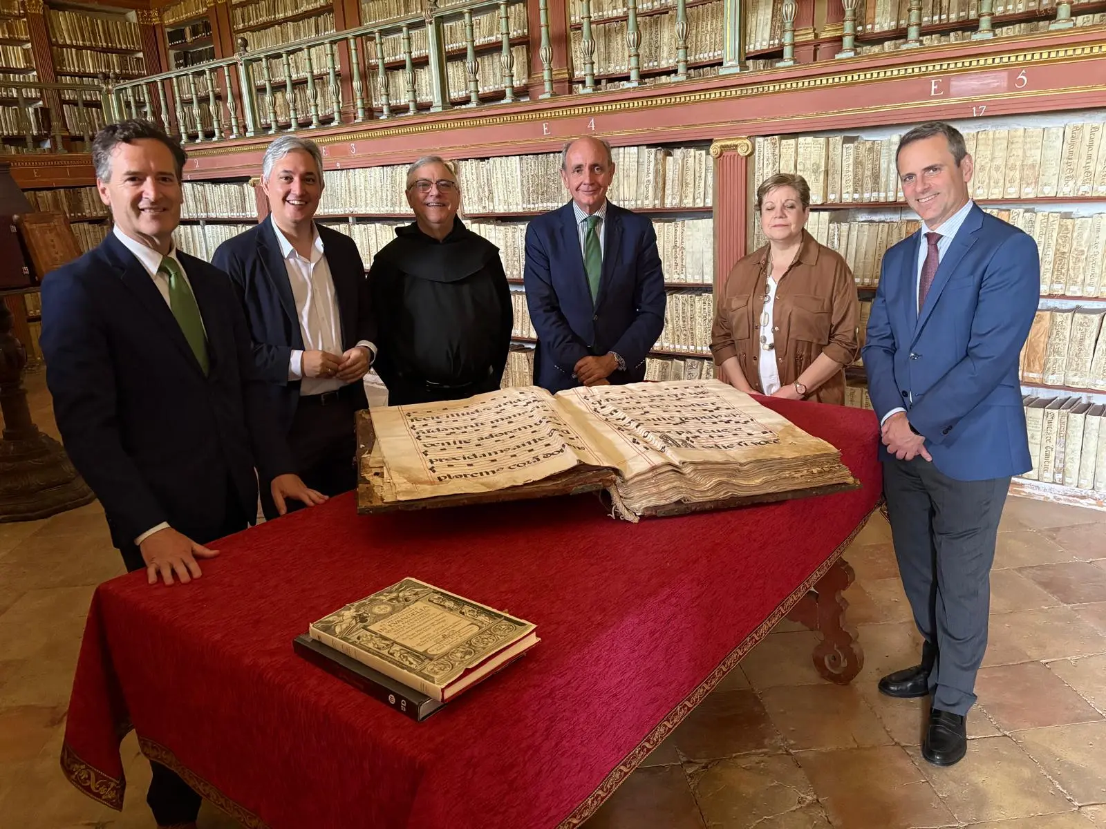 En la imagen posan José Luis Pérez Pastor, vicepresidente de la Fundación San Millán de la Cogolla; Almudena Martínez, coordinadora general de la fundación; Ramón Castresana, director de la Fundación Iberdrola España; Antonio Collada, responsable de arte, cultura y biodiversidad; José Ramón Pérez, prior del Monasterio de Yuso; y Eduardo Ryan, delegado institucional de Iberdrola en La Rioja, en una biblioteca histórica junto a un manuscrito antiguo expuesto sobre una mesa.
