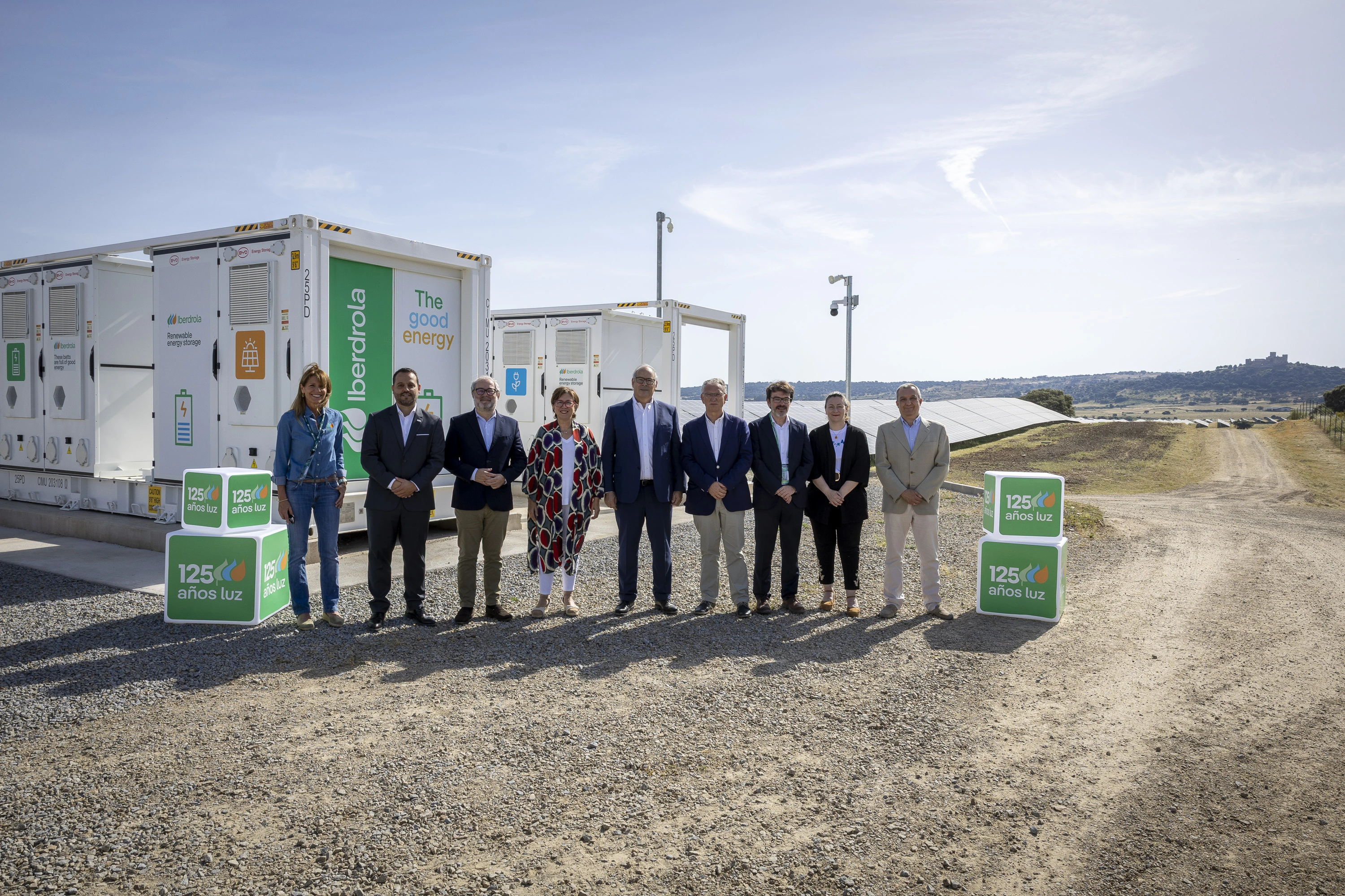 Grupo de personas de pie frente a contenedores de almacenamiento de energía renovable de Iberdrola, con paneles solares y paisaje abierto al fondo.