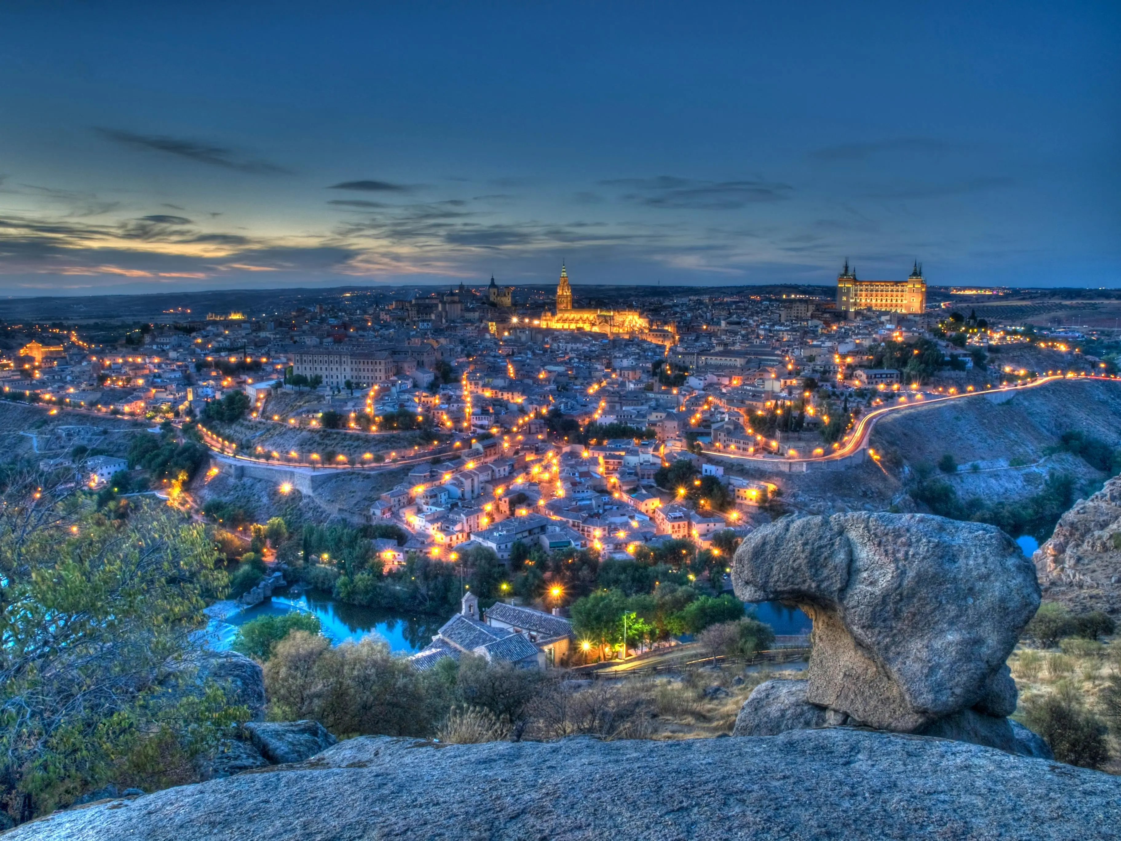 Vista panorámica nocturna de Toledo con luces encendidas, mostrando edificios históricos y la ciudad sobre colinas al atardecer.