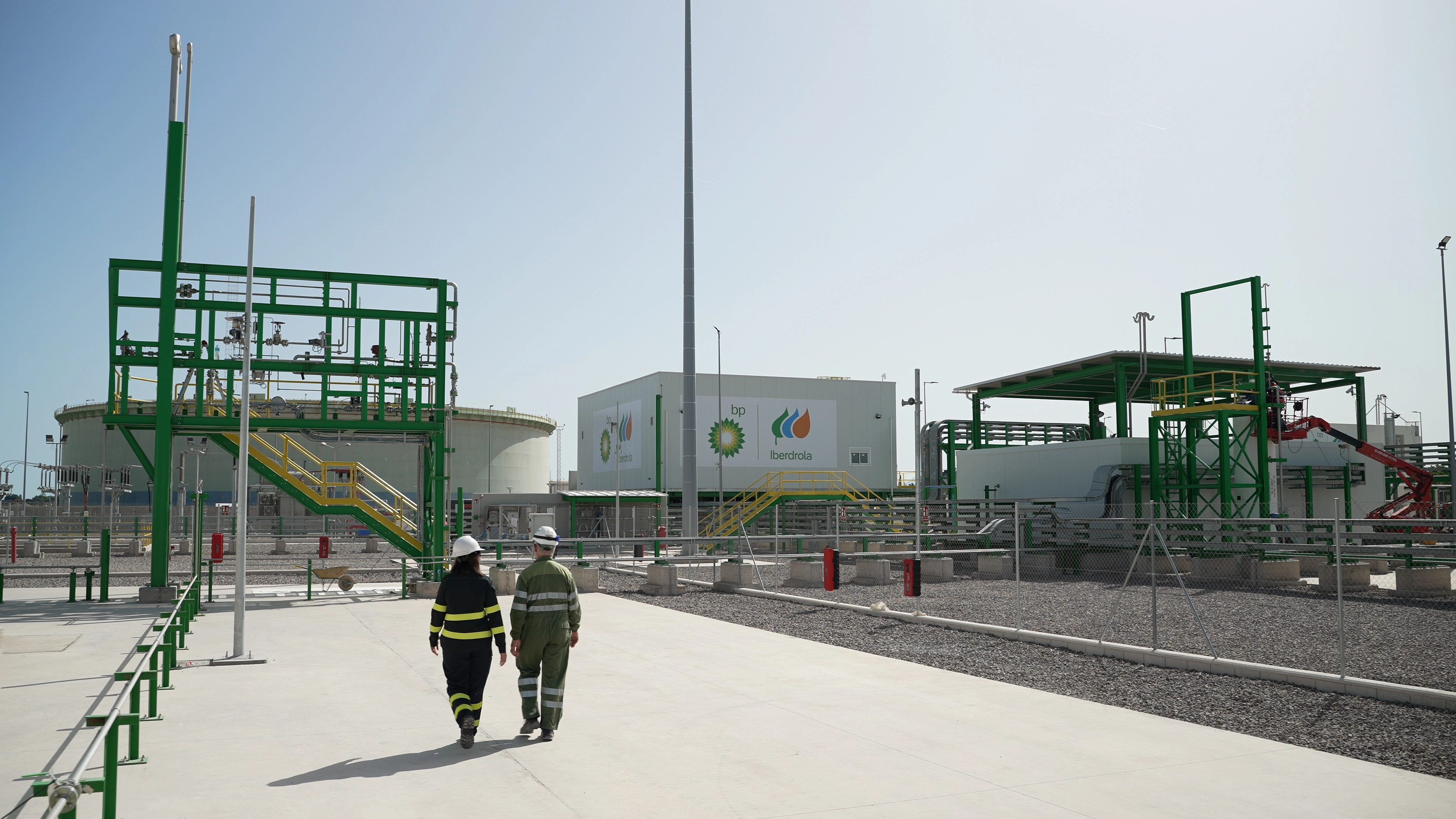 Dos trabajadores con equipos de protección en planta de hidrógeno verde con instalaciones industriales, tuberías y tanques.