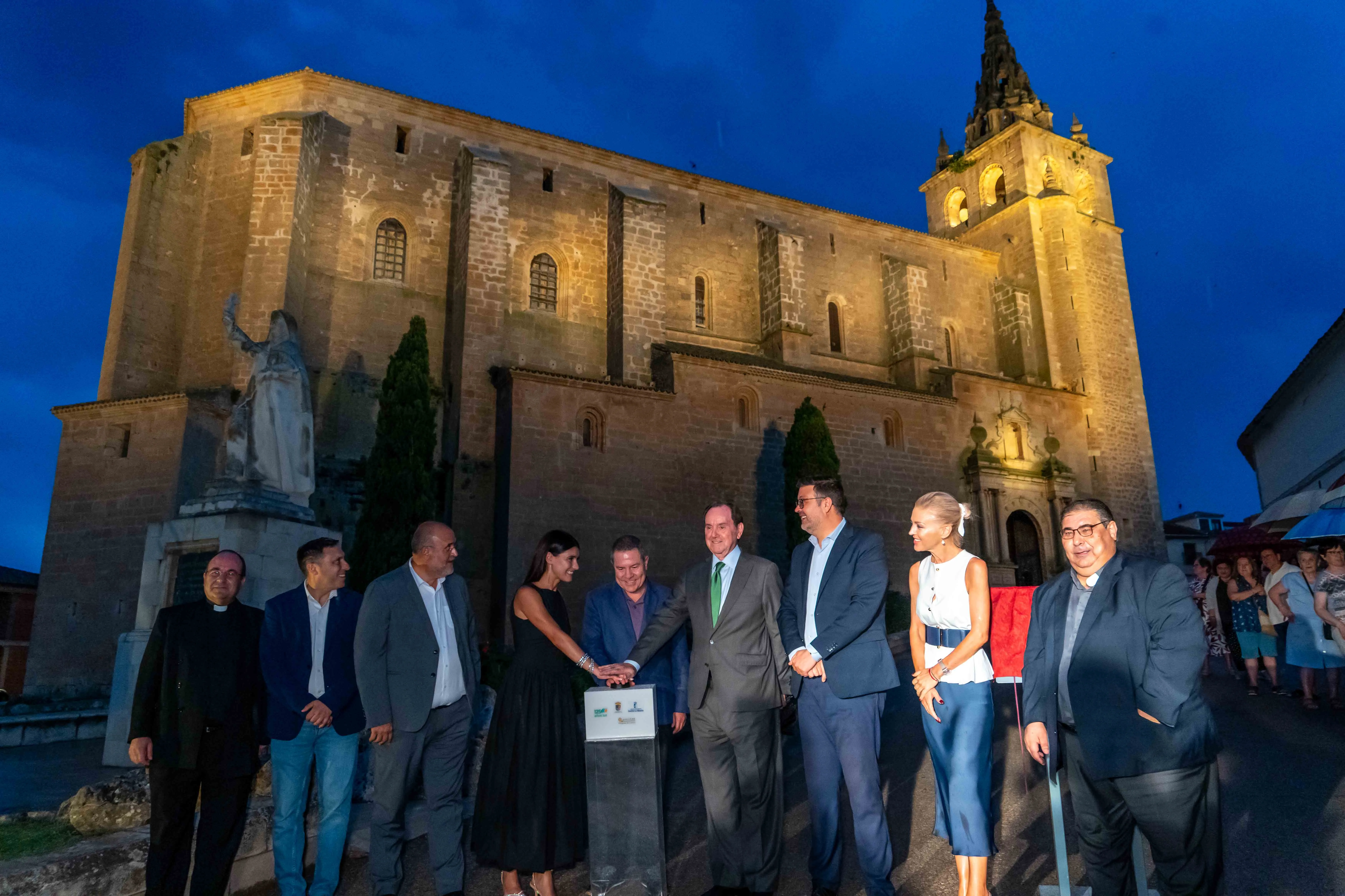Inauguración de iluminación artística en una basílica histórica con representantes y asistentes frente al edificio iluminado.