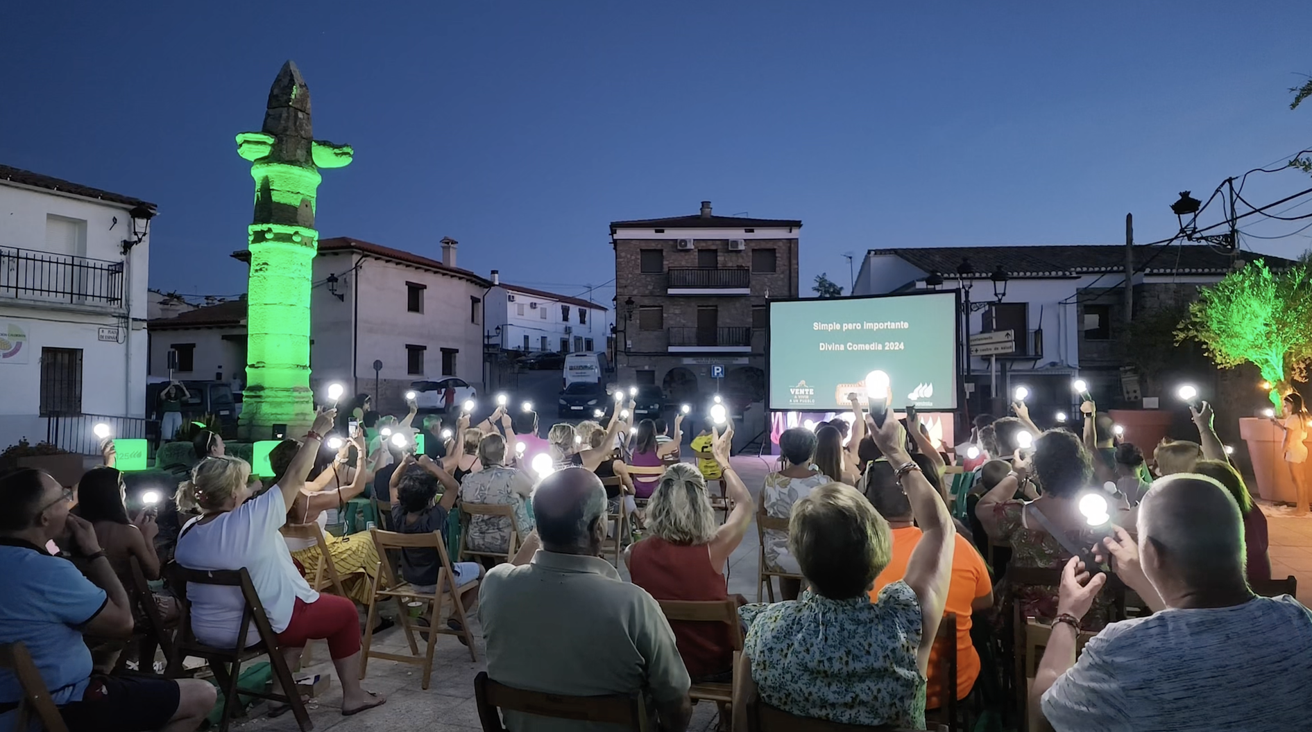 Asistentes reunidos en una plaza durante la proyección de la película “Divina Comedia 2024” en el Festival de Microcine de Verano, sosteniendo luces portátiles mientras cae la noche.