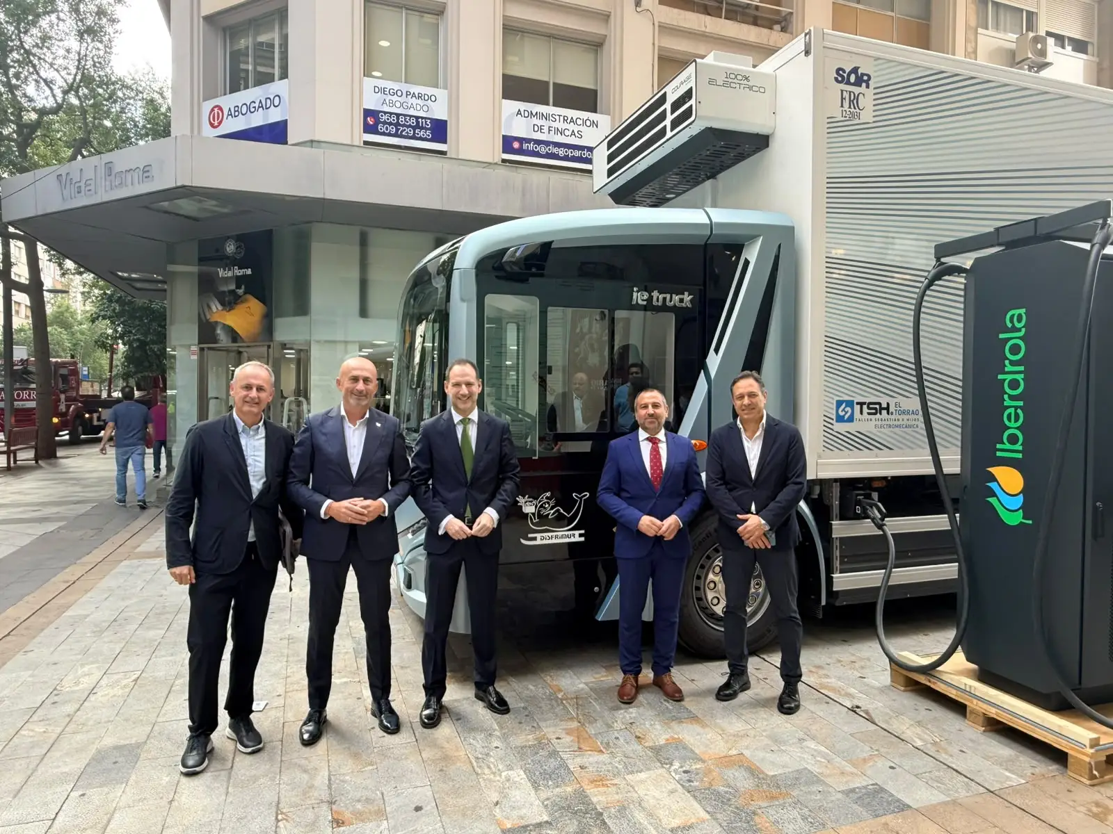 Cinco representantes posan junto a un camión 100 % eléctrico y un punto de recarga de Iberdrola durante una jornada sobre electrificación del transporte.