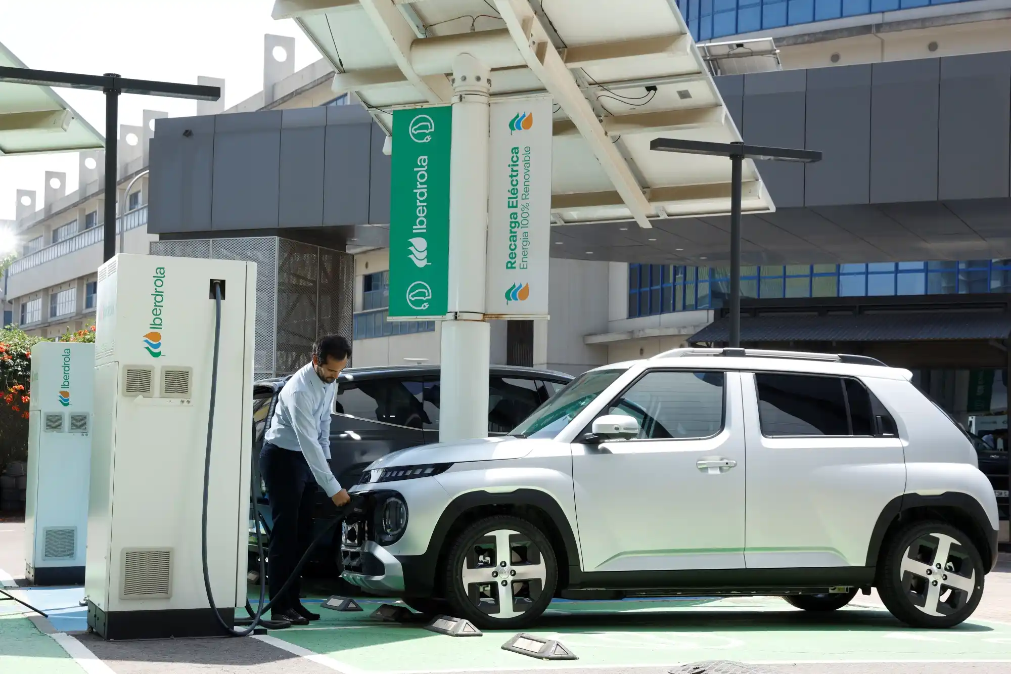 Un hombre conecta un vehículo eléctrico blanco a un punto de recarga de Iberdrola, alimentado con energía 100 % renovable, en un aparcamiento exterior.