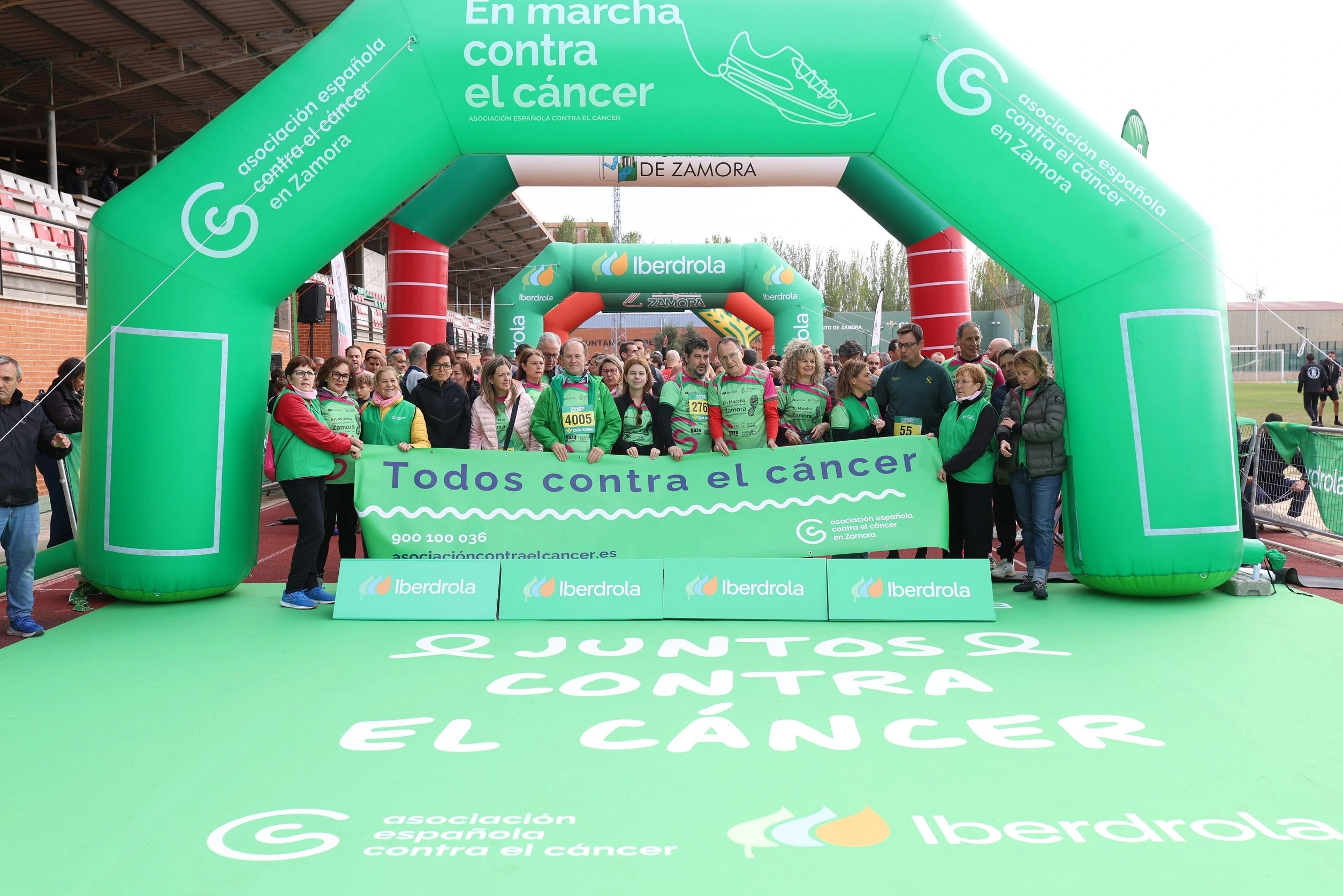Participantes en la marcha solidaria «En marcha contra el cáncer» en Zamora posan con una pancarta de «Todos contra el cáncer» bajo un arco verde de la Asociación Española Contra el Cáncer, con el patrocinio de Iberdrola.
