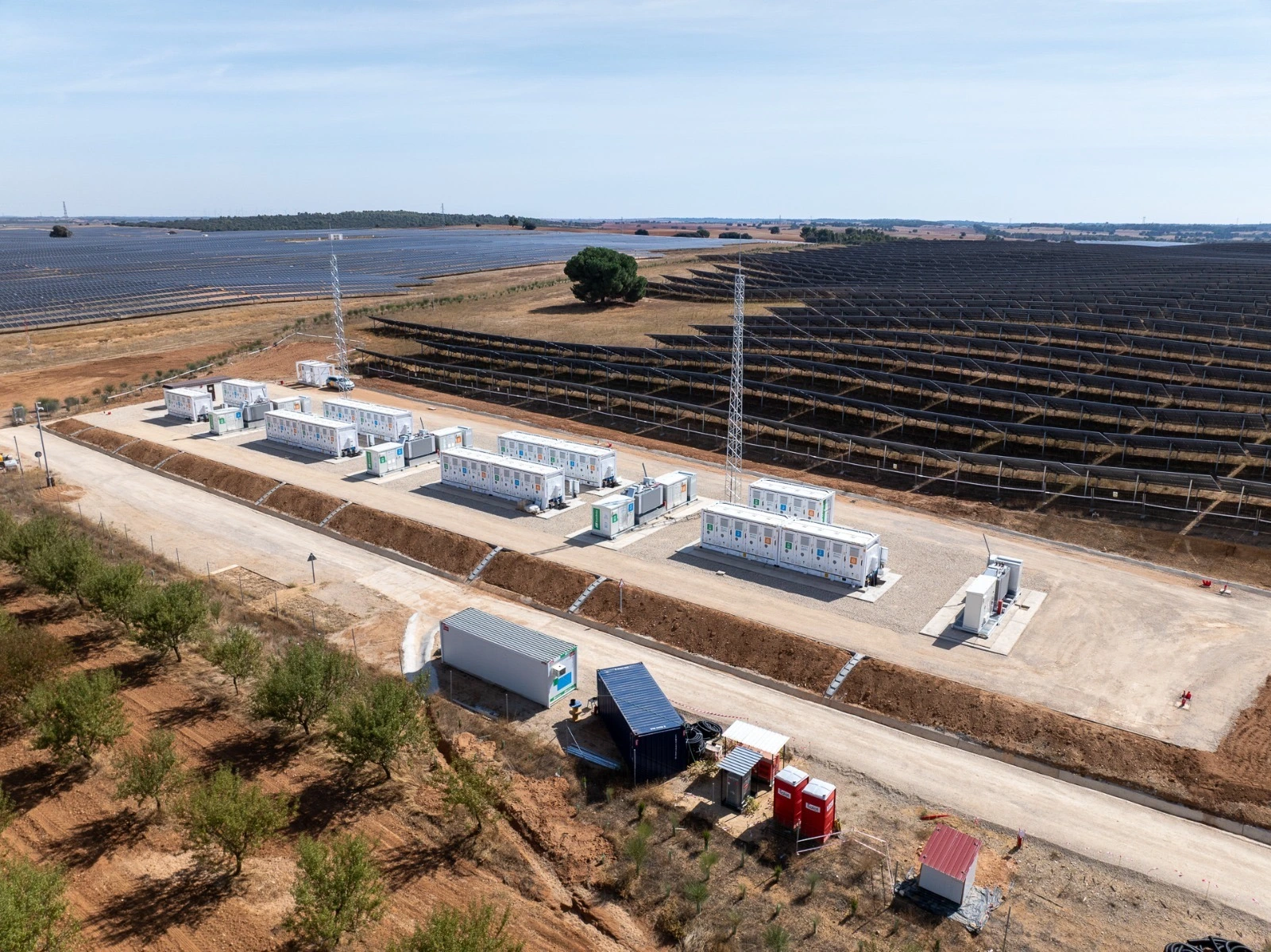 Vista aérea de una planta de baterías de almacenamiento de energía, con varios contenedores blancos alineados en el suelo. En el fondo, se pueden ver los paneles solares de un campo fotovoltaico.