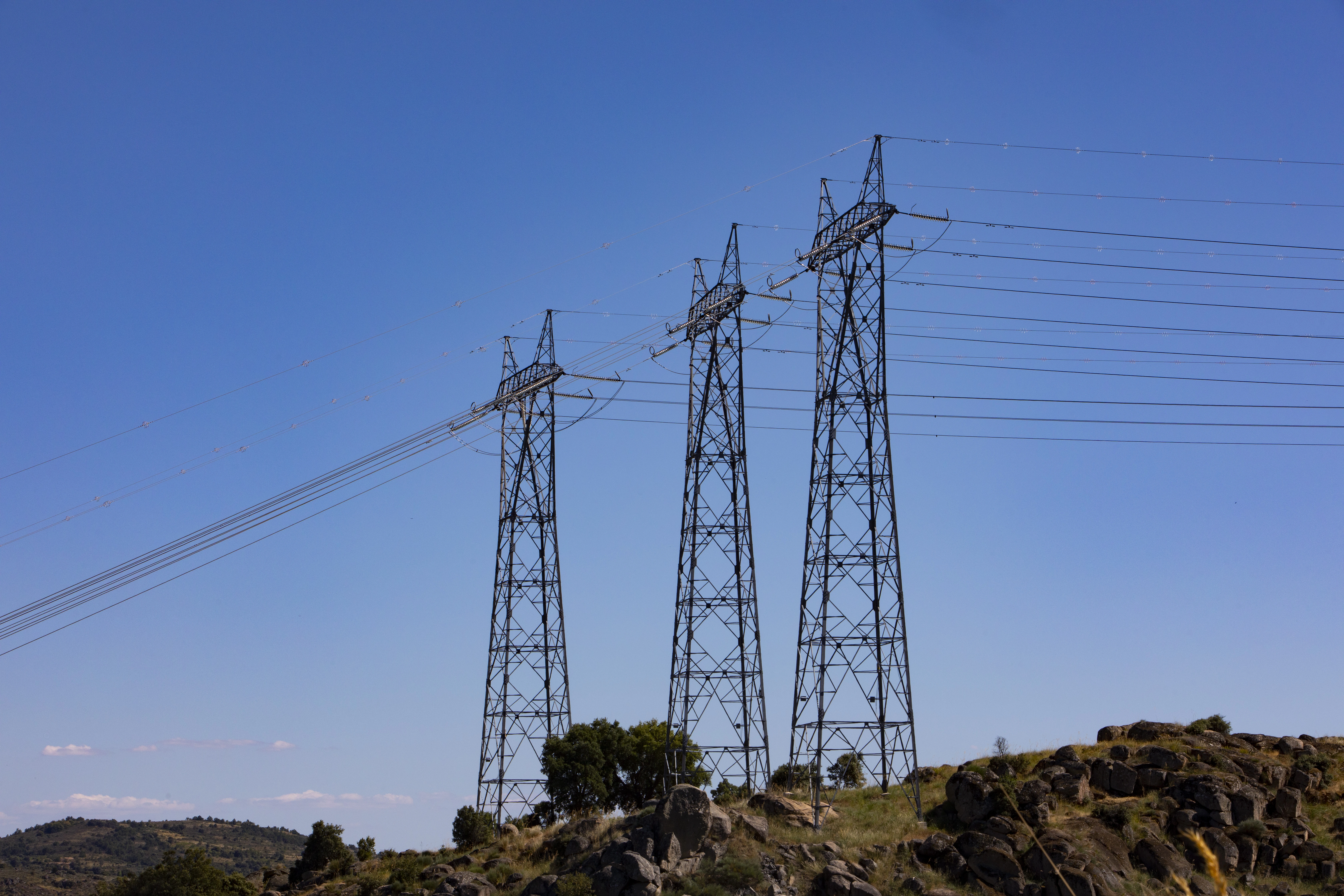 Tres torres eléctricas situadas sobre un monte verde con arbustos bajo un cielo azul despejado