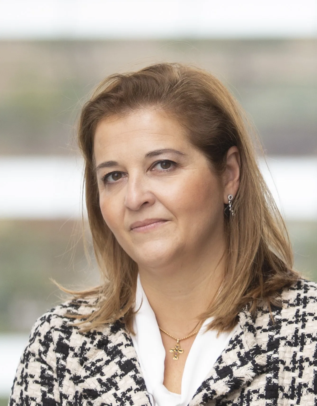 Retrato profesional de Eva Mancera, CEO de i-DE Redes Eléctricas Inteligentes. Viste una chaqueta con patrón de tweed en blanco y negro sobre una blusa blanca, y lleva un collar con una pequeña cruz dorada. El fondo está desenfocado, sugiriendo un entorno de oficina luminoso